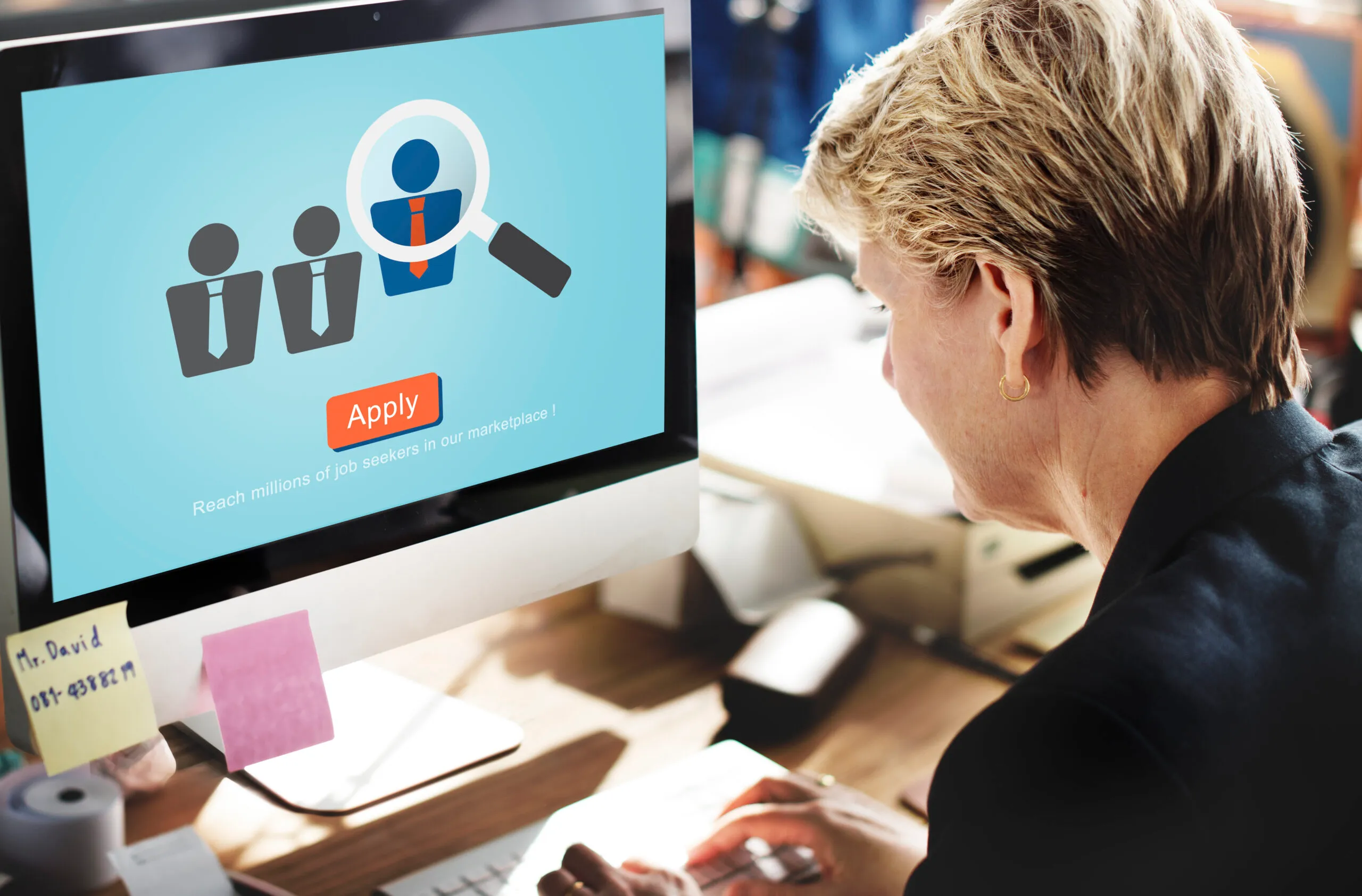 A woman at a desktop computer viewing a candidate-search graphic with an Apply button