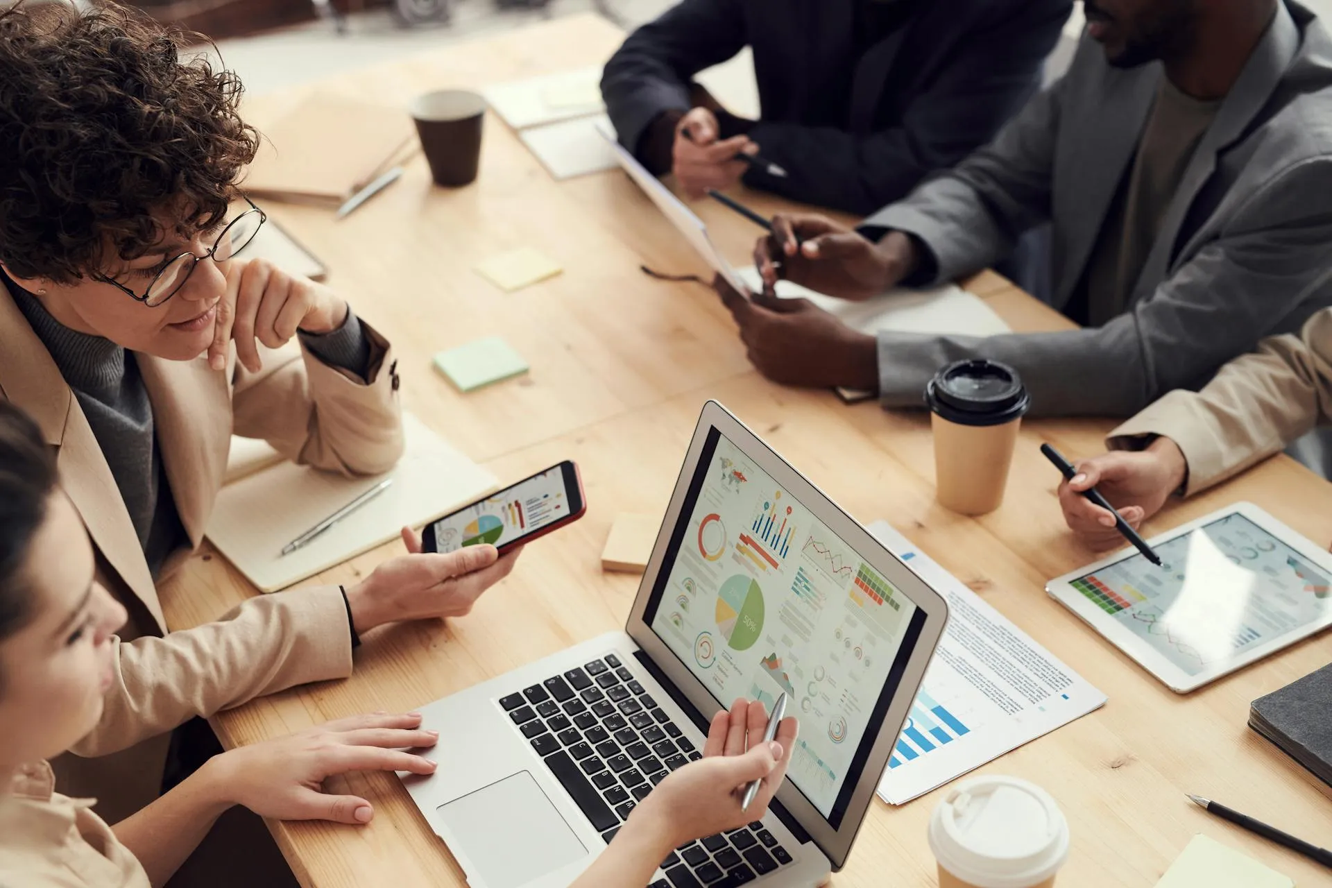 A team reviewing charts on a laptop, phone, and tablet around a table with coffee cups