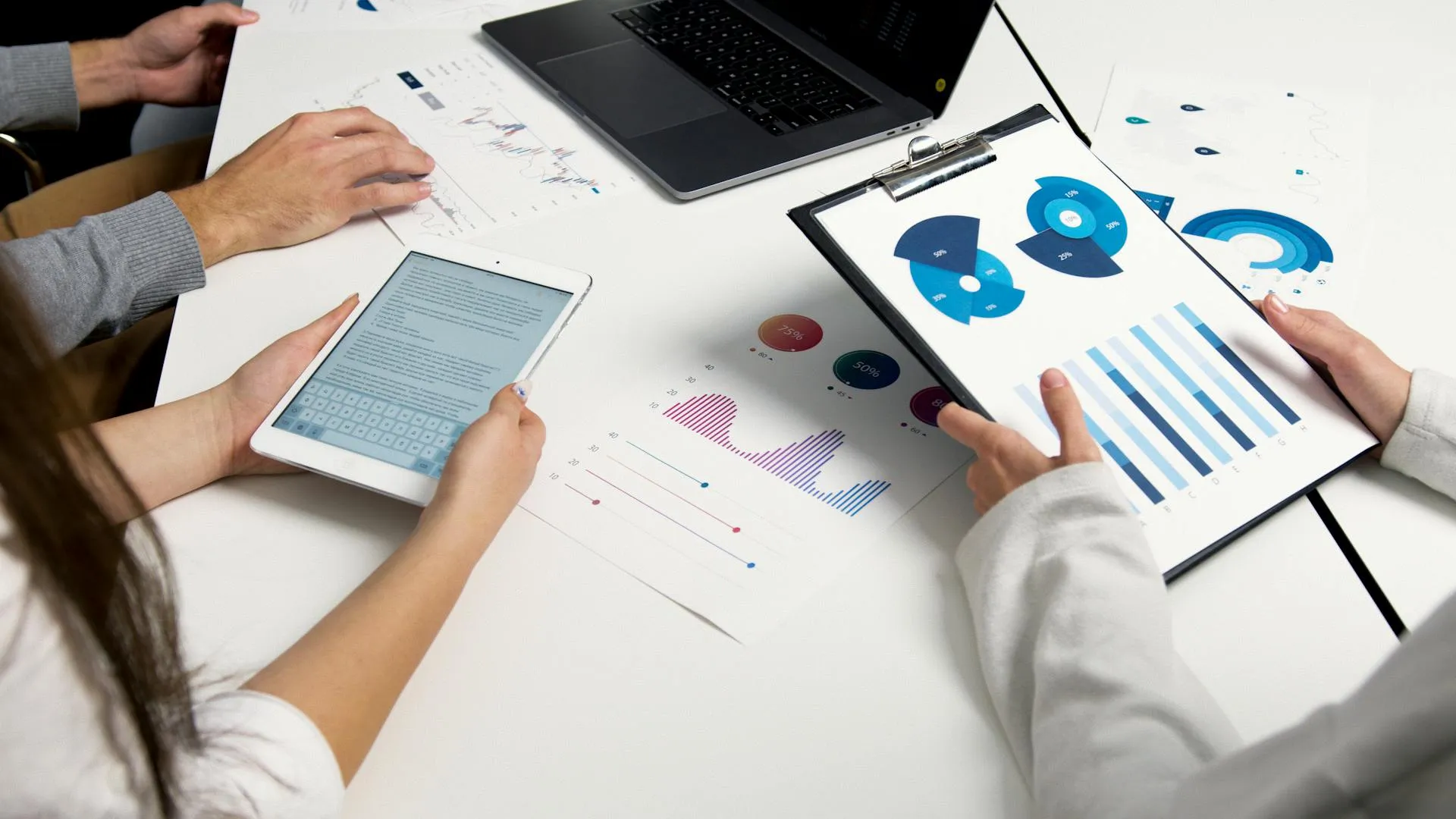 Colleagues reviewing printed pie and bar charts on a clipboard next to a tablet and laptop